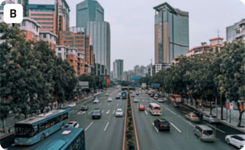 B. Fotografia de uma avenida movimentada em uma cidade grande. Alguns veículos que estão na avenida possuem retângulos inseridos digitalmente, envolvendo-os. Há carros e ônibus, muitas árvores nos canteiros laterais, e arbustos no canteiro central, assim como edifícios muito altos ao fundo. O céu está cinza.