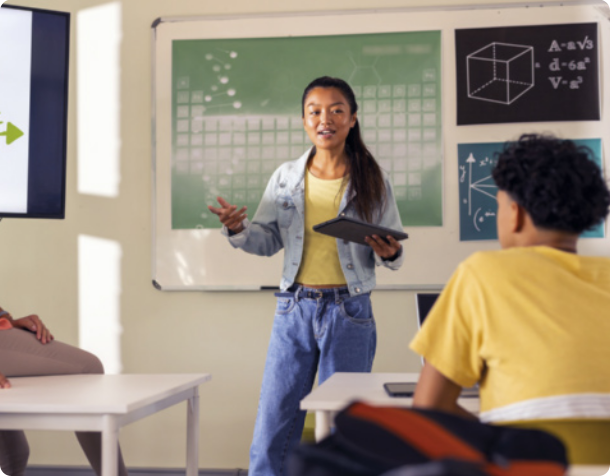 Fotografia de uma jovem fazendo uma apresentação em sala de aula. A jovem está falando, gesticulando com uma das mãos, enquanto segura um 'tablet' com a outra. Ela está usando os cabelos presos, uma camiseta, jaqueta e calça. Atrás da jovem há um quadro com uma imagem da tabela periódica, uma imagem de cubo, algumas fórmulas matemáticas, e um gráfico. Ao lado da jovem há uma janela aberta, e à sua frente há carteiras escolares. Uma das carteiras está ocupada por um jovem, que está assistindo à apresentação, e para a qual a jovem está direcionando o seu olhar.