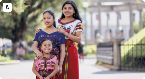 A. Fotografia de três jovens de traços indígenas sorrindo em um parque. As jovens estão dispostas uma atrás da outra, conforme a idade: a mais jovem na frente, e a mais velha atrás. A mais jovem é uma criança. Todas estão usando roupas tradicionais coloridas com cores vibrantes e bordados. Ao fundo, está o parque, com algumas árvores, pessoas, gramados, e uma construção clássica com colunas brancas.