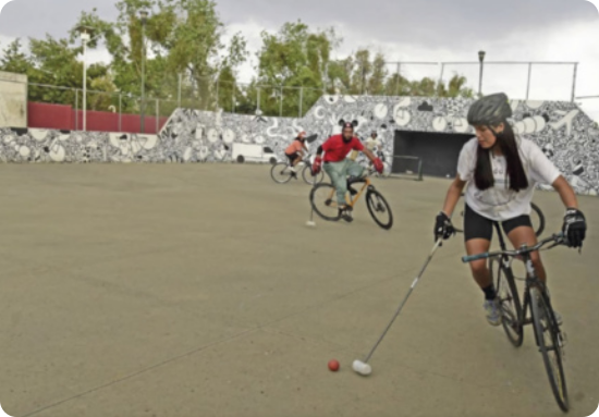 Fotografia de dois jovens jogando 'bike' polo em uma quadra de esportes. À frente na foto está uma jovem pedalando uma bicicleta e segurando um taco com uma das mãos e o guidão da bicicleta com a outra. Com o taco, a jovem move uma bola pequena no chão. A jovem usa capacete e luvas, uma camiseta e bermuda. Mais ao fundo há um jovem pedalando uma bicicleta, também segurando um taco e o guidão da bicicleta, porém ele não está em posse da bola. O jovem usa camiseta, calça e luvas. Mais ao fundo há outra pessoa pedalando uma bicicleta, um muro desenhado, uma cerca acima do muro, e muitas árvores que estão do lado de fora da quadra.