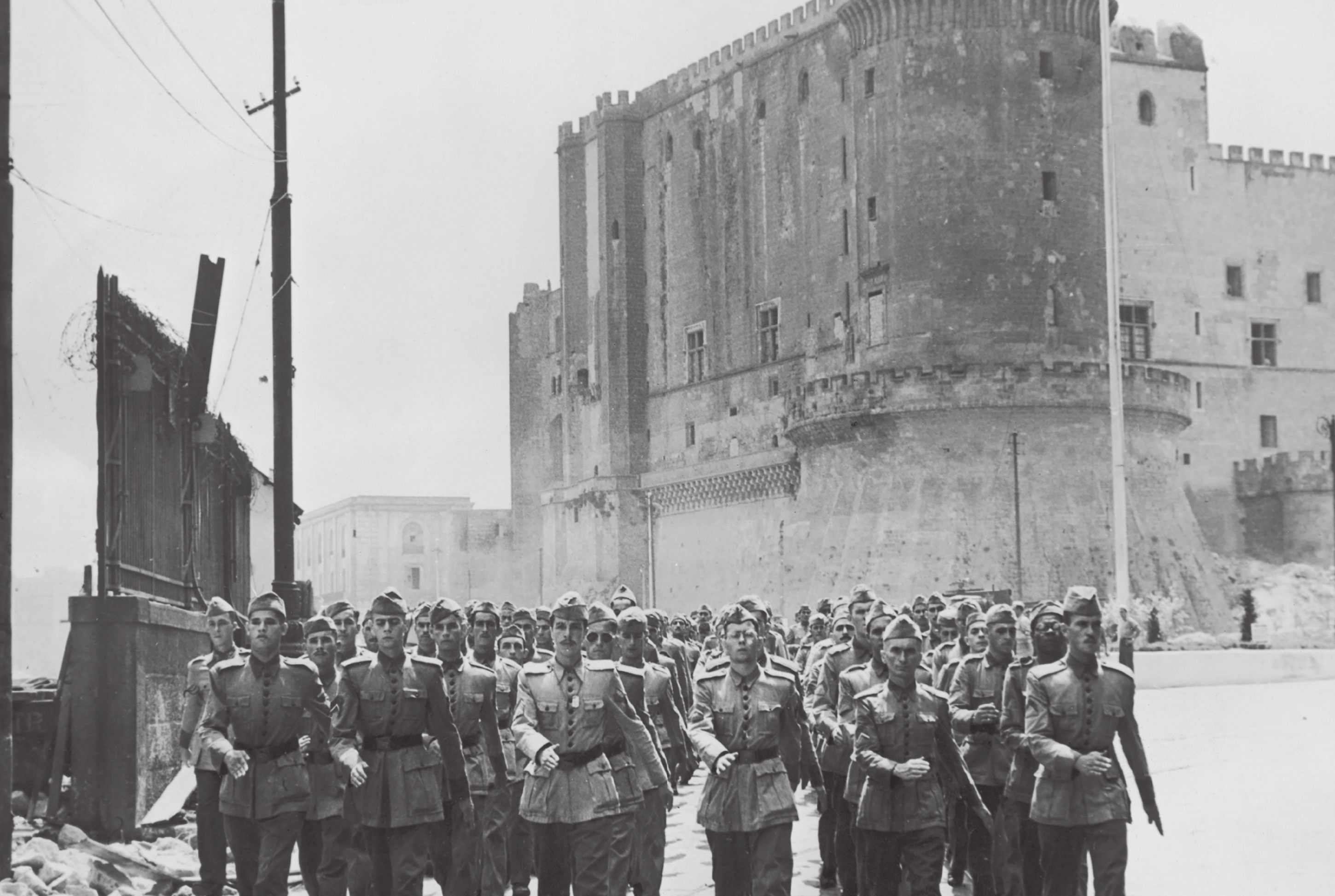 Fotografia em preto e branco de soldados expedicionários brasileiros em Nápoles. Na imagem, muitos soldados juntos marcham pela rua. Eles usam uniforme, farda militar e  quepes na cabeça.