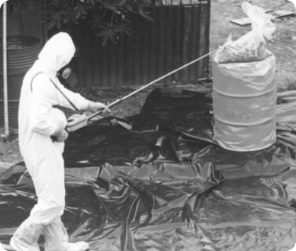 Fotografia em preto e branco de um homem usando macacão de proteção , com capuz, máscara e botas, segura um equipamento, perto de uma cápsula. O chão está forrado com proteção.