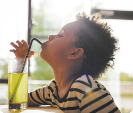 Fotografia de um menino, tomando suco, em um copo, com um canudo, que dobra a ponta. O menino usa cabelo curto crespo e camiseta listrada.