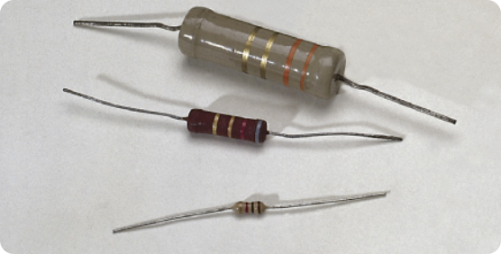 Fotografia de três resistores de carvão, de tamanhos diferentes enfileirados do maior acima da para o menor, abaixo. Os resistores são objetos cilíndricos de metal com dois fios saindo em cada extremidade.