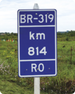 Fotografia de uma placa retangular de rodovia, escrito: 'BR-319. Quilômetro 814. RO'. Ao fundo, há uma área com vegetação de gramíneas.