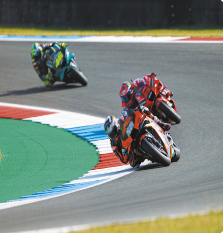 Fotografia de motociclistas em motos, inclinados, na curva da pista. Eles usam uniformes e capacetes de cores diferentes.