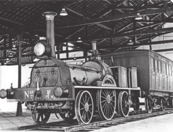 Fotografia em preto branco, de uma locomotiva antiga. Ela tem a frente com uma chaminé e um farol, três rodas de cada lado e está puxando um vagão. Acima um telhado de um galpão.
