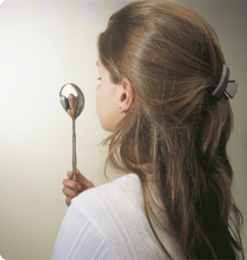 Fotografia de uma mulher de cabelos longos , segurando uma colher na mão. Ela observa seu reflexo sendo refletido na colher.