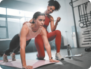 Fotografia de duas mulheres fazendo exercício físico. A aluna se equilibra em um tapete, apoiada nos braços e pernas. enquanto a professora, está ao seu lado ajoelhada em uma perna. ao fundo equipamentos de ginástica.