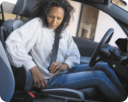 Fotografia de uma mulher, dentro de um veículo, colocando o cinto de segurança. Ela está sentada no banco do carro, usa blusa de manga longa e calça comprida. Tem o cabelo crespo, na altura do ombro.