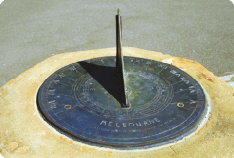 Fotografia de um relógio de Sol. Ele é formado por uma pedra circular com marcações, colocado horizontalmente sobre uma rocha elevada. Em seu centro, há uma segunda peça triangular colocada na vertical, que produz uma sombra que aponta para as marcações.