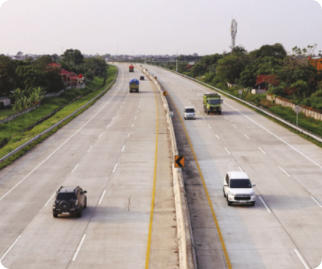 Fotografia de uma rodovia de dois sentidos. Carros e caminhões trafegam nos dois sentidos da via. Ao centro da rodovia, há um pequeno muro separando as pistas. De ambos os lados da estrada, há vegetação ao longo da margem.
