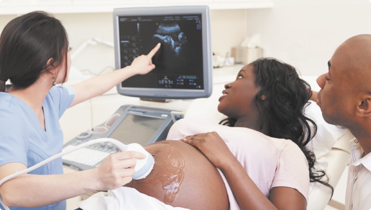 Fotografia de uma mulher grávida, deitada em uma cama de hospital, com a barriga despida, ela observa em uma tela de computador, a imagem de um ultrassom, que está sendo passado por sua barriga, por uma outra mulher que usa uniforme e aponta para a tela. Ao lado um homem, observa a tela.