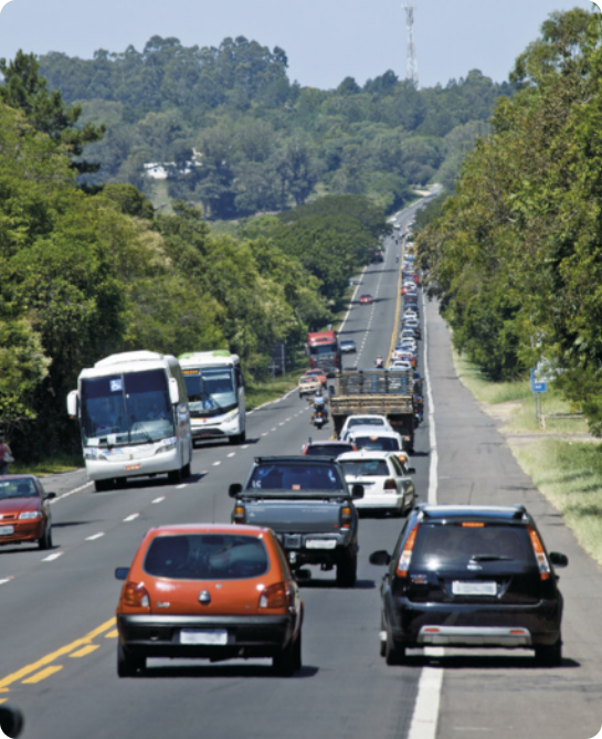 Fotografia de uma rodovia de mão dupla, com vários carros. Um dos carros da pista da direita trafega pelo acostamento. Ao fundo há vegetação.