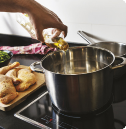 Imagem de uma panela de metal, em cima de uma superfície plana de um fogão por indução. Na imagem, uma mão de uma pessoa, segura uma garrafa de óleo de cozinha. Imagem de alimentos ao lado da panela.