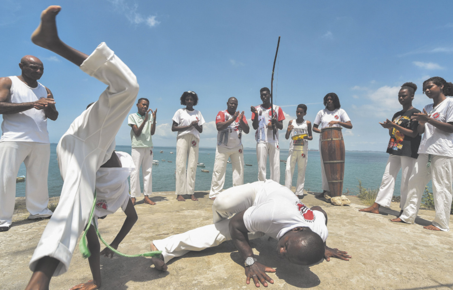 Fotografia de pessoas jogando capoeira ao ar livre. Na imagem, duas pessoas estão ao centro ,um coma as pernas levantadas, apoiando as mãos no solo. O outro está abaixado no chão enquanto várias pessoas assistem.