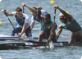 Fotografia de pessoas praticando canoagem. Eles estão cada um em uma canoa, dentro da água, usam um remo, cada, estão com roupa esportiva e de toca de borracha.