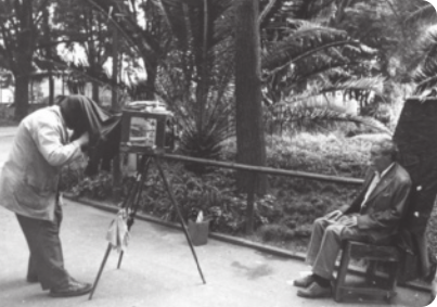 Fotografia em preto e branco, de um fotógrafo, tirando o retrato de um homem sentado. A câmera fotográfica, tem formato de uma caixa, apoiada em hastes .O fotógrafo está com a cabeça tampada por um pano, preso na câmera.