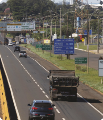 Fotografia de uma rodovia com trânsito de veículos. Um carro e um caminhão aparecem em primeiro plano, o carro na faixa da esquerda e o caminhão na faixa da direita. Eles estão a alguns metros de distância entre si.