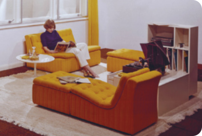 Fotografia de uma sala decorada com móveis antigos. Uma pessoa está sentada em uma poltrona, com uma mesinha circular ao lado. Uma televisão pequena em cima de um móvel, ao lado uma estante e um divã.