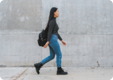 Fotografia de uma mulher andando na rua, ela tem cabelos longos, usa blusa de manga longa e calça comprida, tem uma bolsa pendurada no ombro.