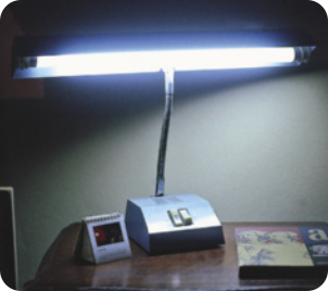 Fotografia de uma luminária de mesa, acesa, em cima de uma mesa. A luminária é em formato cilíndrico horizontal.