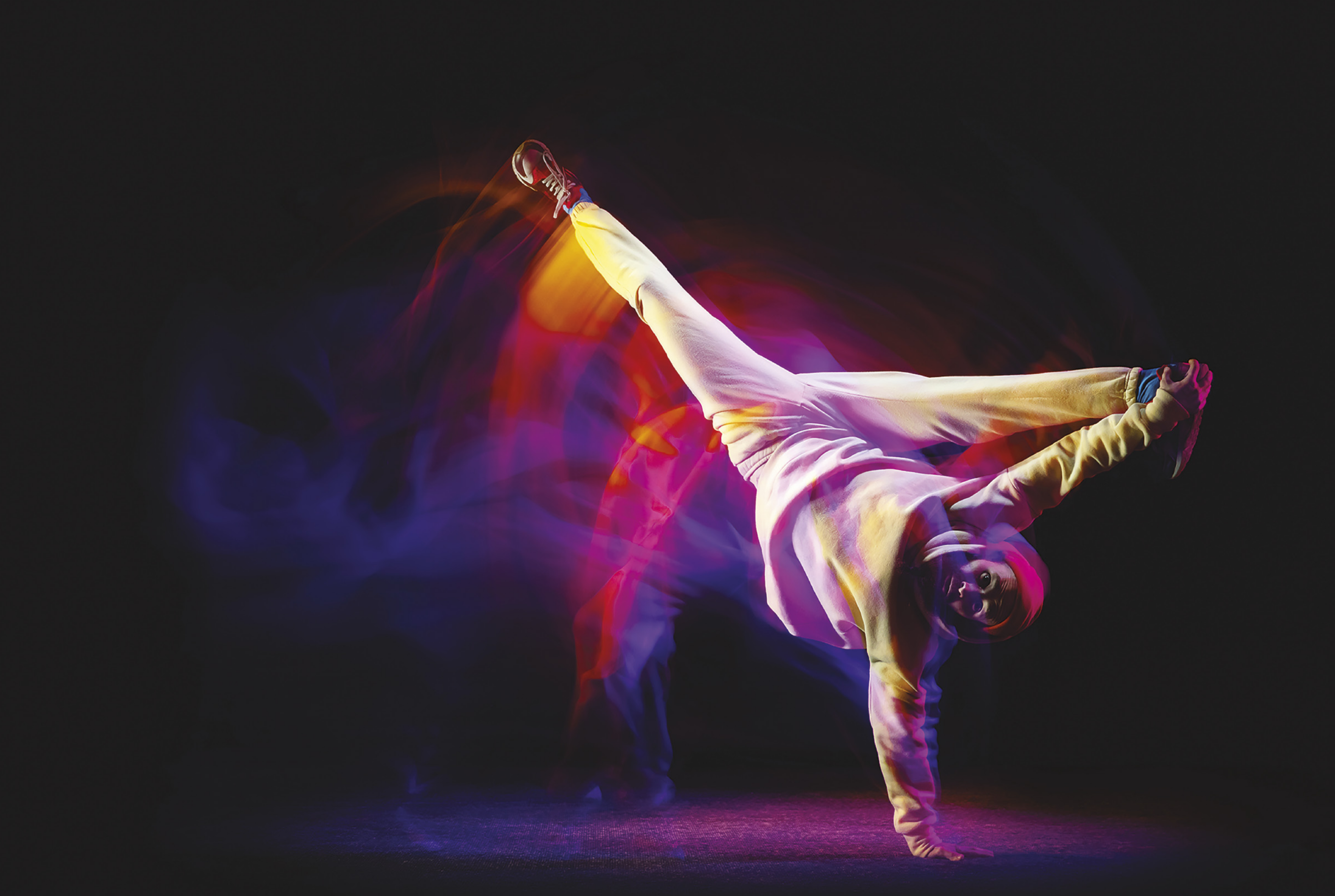 Fotografia de uma pessoa realizando um passo de dança. Ela está se equilibrando em uma das mãos, com a palma apoiada no chão, enquanto as pernas estão abertas, direcionadas para cima. O outro braço está esticado para frente.