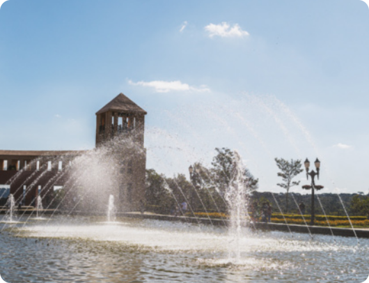Fotografia da fonte do Parque Tanguá. Em primeiro plano, jatos de água da fonte se elevam em diferentes alturas sobre um espelho d’água, formando arcos. Ao fundo, vê-se uma estrutura de alvenaria e árvores.