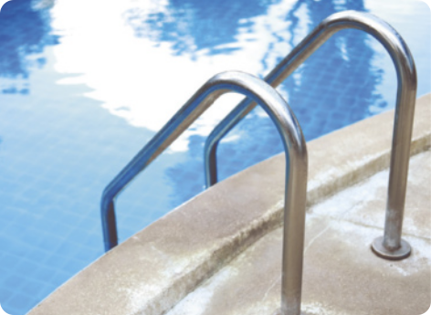 Fotografia de uma escada de piscina. Duas barras de metal curvadas paralelas, fixadas no chão. Na imagem o céu está refletindo na água da piscina.