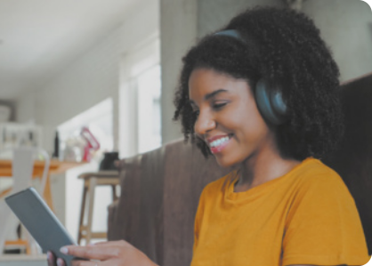 Fotografia de uma jovem usando fones de ouvindo, olhando para um celular. Ela usa cabelo crespo e está sorrindo.