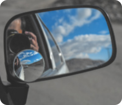 Fotografia de um espelho retrovisor de um carro, refletindo a imagem da pessoa  que conduz o carro, uma parte da lateral do carro e o céu.