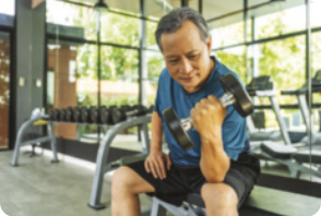 Fotografia de um homem praticando musculação em uma sala de academia. Ele segura um peso em uma das mãos, está sentado, usa cabelo curto crespo, roupa esportiva, camiseta e shorts. Ao fundo imagem de pesos enfileirados.