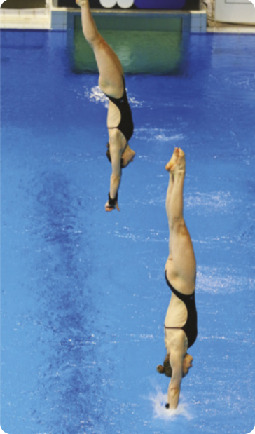 Fotografia de duas mulheres mergulhando em uma piscina, lado a lado. Ambas vestem maiôs pretos e estão com a cabeça posicionada entre os braços, que estão estendidos para baixo, em direção à superfície da piscina. Uma das mulheres já está com um dos braços parcialmente submerso na água, enquanto a outra se encontra ainda próxima à superfície, prestes a entrar.