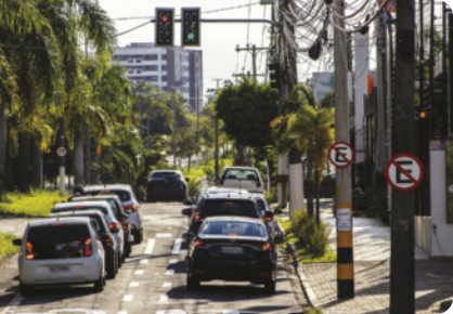 Fotografia de carros parados em um semáforo, com os faróis dos freios ligados. Na imagem aparecem árvores, postes de luz e placas de trânsito.