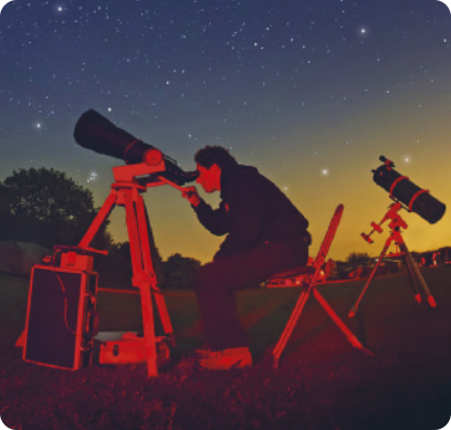 Fotografia de um homem, sentado em uma cadeira ao ar livre, observando o céu a noite, por um telescópio.