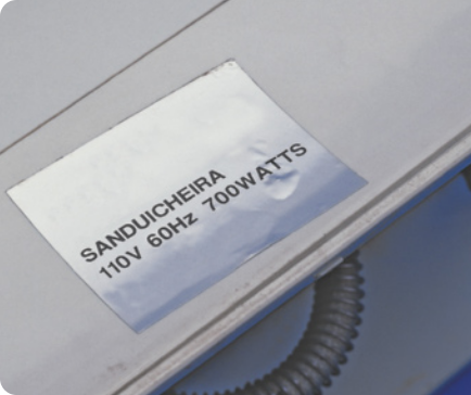 Fotografia de uma etiqueta com especificações técnicas. Na etiqueta está escrito 'SANDUICHEIRA',' 110volts',' 60 hertz' e '7000 watts.'