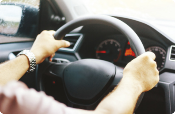 Fotografia de uma pessoa segurando o volante de um carro, com as duas mãos. Na imagem aparece o interior do veículo, o velocímetro e o espelho retrovisor.