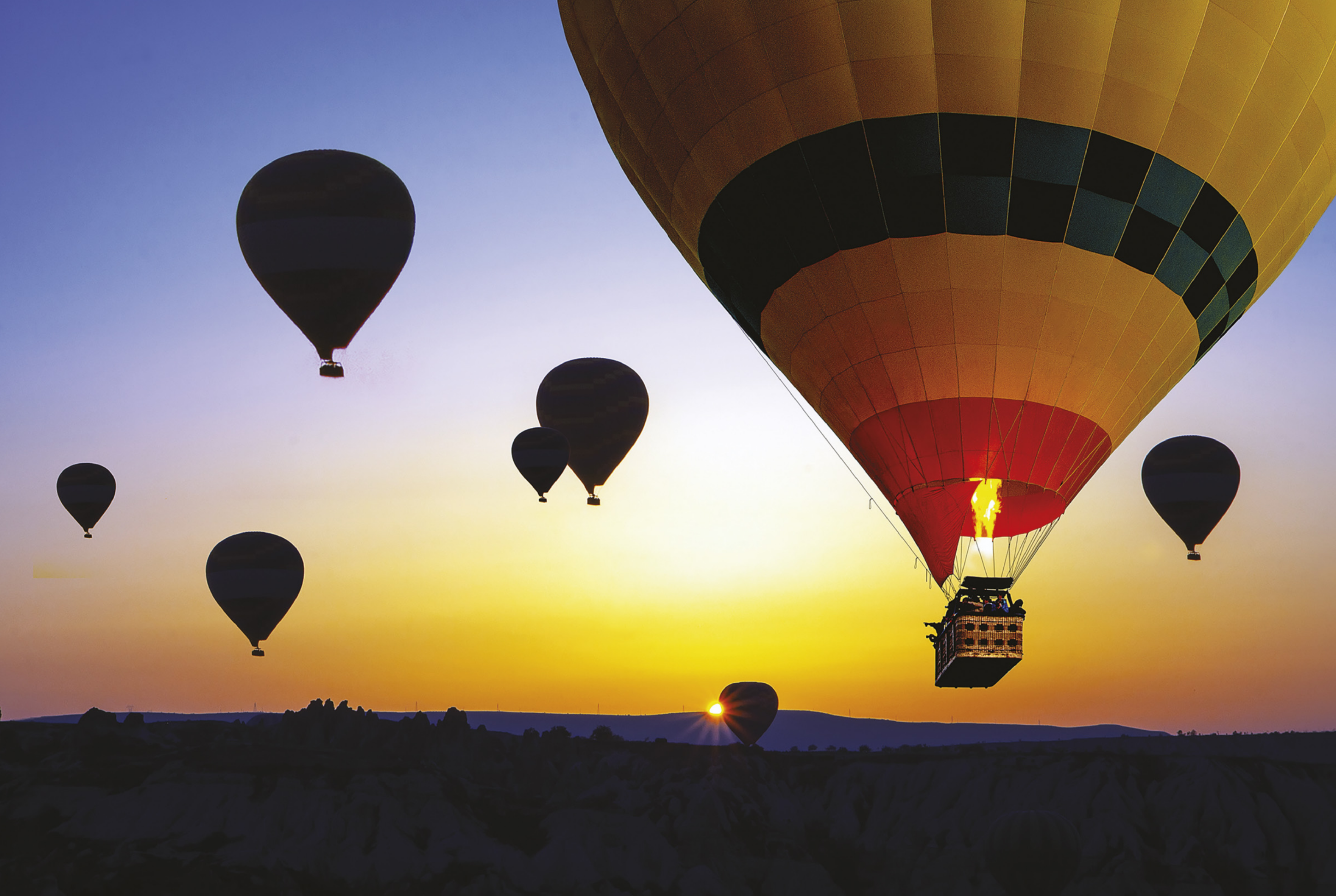 Fotografia de vários balões flutuando no céu, ao fundo o Sol se pondo. Um balão está em destaque, colorido. Ele tem  fogo saindo por baixo e uma cesta pendurada.