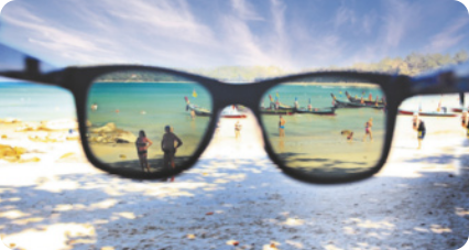 Fotografia de óculos de Sol, lentes quadradas. Ao fundo paisagem de uma praia. Pelas lentes do óculos, vemos parte da paisagem ao fundo em outro tom de luz.