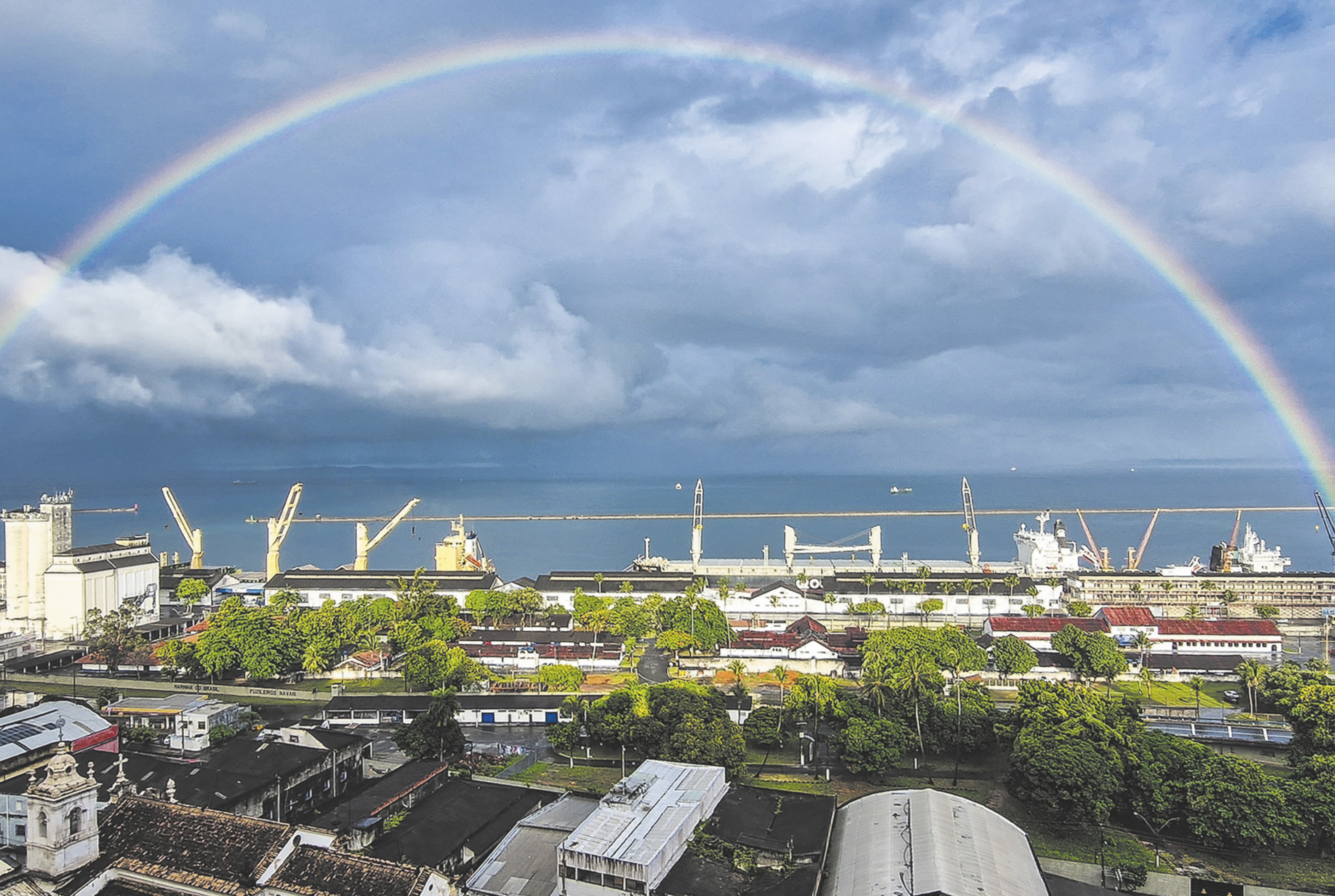 Fotografia de um arco-íris no céu, com nuvens, abaixo telhados.
