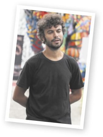 Fotografia de Bordalo II, um homem branco, com barba, bigode e cabelos curtos e ondulados, vestindo uma camiseta preta. Ele está com a boca entreaberta, sugerindo que está falando, e mantém ambos os braços posicionados atrás do corpo.