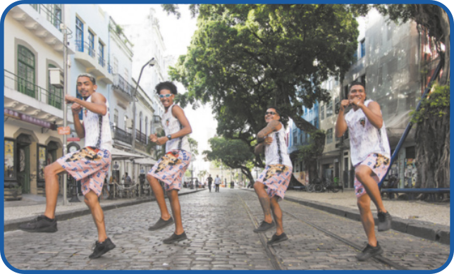 Fotografia de quatro rapazes dançando, sincronizados, em uma rua de paralelepípedos. Eles estão de lado, sorrindo, levantando a perna esquerda levemente flexionada e aproximando os braços do corpo. Todos eles estão usando regata branca com estampas, shorts cor-de-rosa, também estampados, e tênis pretos.