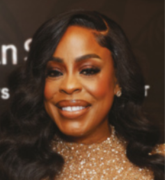 Fotografia de Niecy Nash sorrindo. Mulher de pele negra com cabelos longos e ondulados, penteados para o lado. Ela usa uma blusa dourada, com brilho. O fundo é escuro e desfocado.