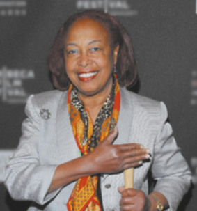 Fotografia de Patrícia Bath sorrindo, segurando o cabo de um martelo com a mão esquerda enquanto tampa a cabeça do martelo com a mão direita. Ela é uma mulher negra de meia-idade, tem cabelos lisos e castanhos na altura dos ombros e está usando um paletó feminino preteado e um lenço estampado no pescoço.