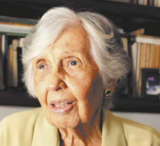 Fotografia de Maria Laura. Ela é uma mulher idosa, de pele branca, cabelos curtos e brancos e usa um casaco amarelo. Ao fundo há uma estante com livros.