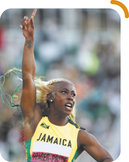 Fotografia de Shelly-Ann Fraser-Pryce. Ela é uma mulher negra, com cabelos compridos tingidos de amarelo e verde, esvoaçantes. Veste uma camiseta amarela com a bandeira da Jamaica e a palavra 'Jamaica' estampada. Está com um dos braços levantados, fazendo o sinal de número um com o dedo indicador, e apresenta uma expressão de vitória no rosto.