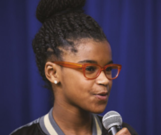 Fotografia de Marley Dias. Ela está com o cabelo preso em um coque alto, usa óculos de armação vermelha e está falando ao microfone. Ao fundo, há uma cortina azul.
