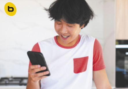B. Fotografia de um jovem sorrindo enquanto olha para a tela do celular que segura com uma das mãos. Ele tem cabelos curtos e lisos, e usa uma camiseta branca com mangas e bolso vermelhos.