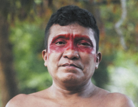 Fotografia de Joseca Yanomami. Ele é um homem indígena, usa o cabelo curto, pintura vermelha nos olhos e está sem camisa.