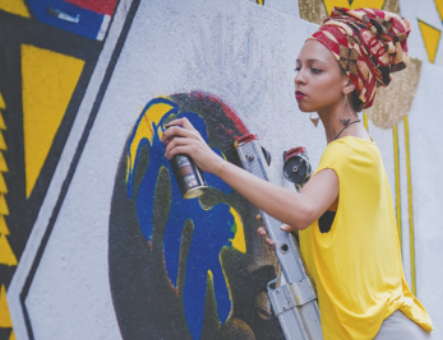 Fotografia de Criola. Ela é uma mulher de pele branca e cabelos presos com um lenço estampado. Usa uma blusa amarela e está aplicando tinta spray em um mural com desenhos coloridos.